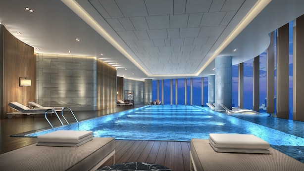 Una piscina interior moderna con un muelle de madera, sillones de descanso y vistas al cielo a través de grandes ventanales.