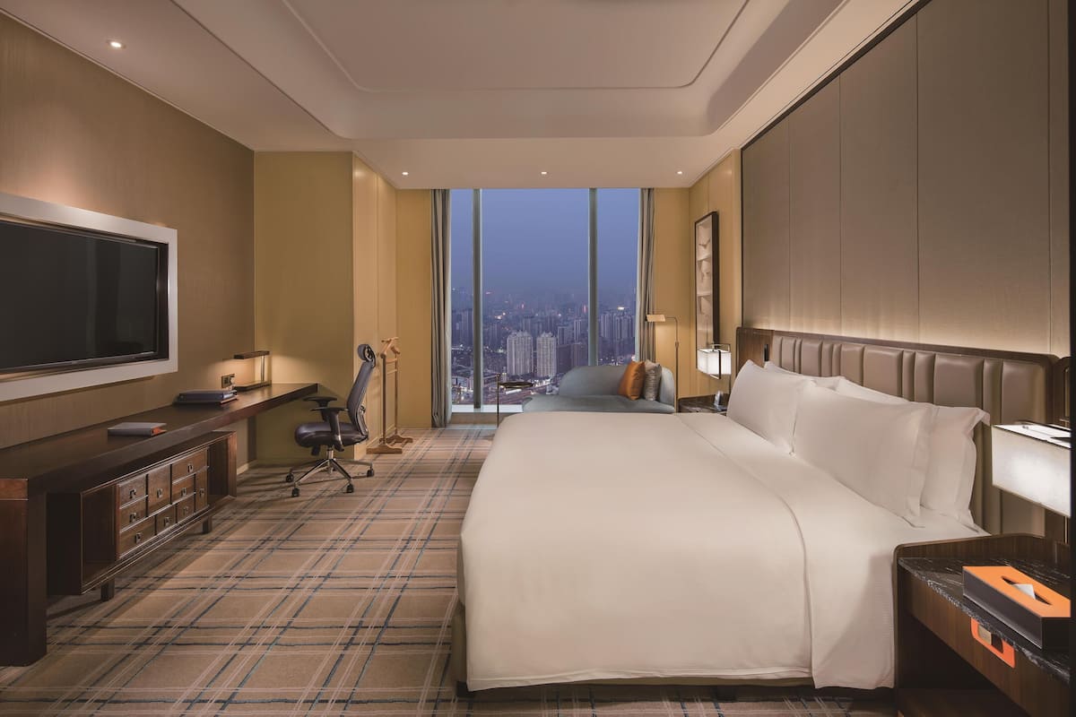 A hotel room with a large bed, a desk with a flat-screen TV, a chair, a desk lamp, and a view of the city through large windows.