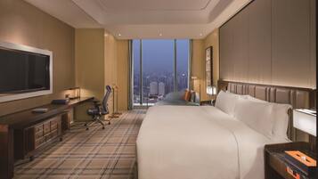 Habitación de hotel con una cama grande, un escritorio con televisor de pantalla plana, una silla, una lámpara de escritorio y vistas a la ciudad a través de amplias ventanas.