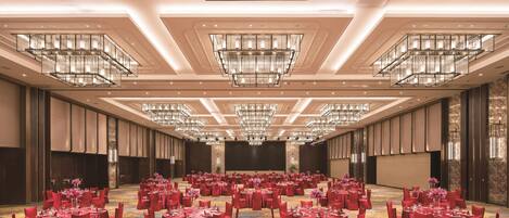 Un amplio salón de banquetes con varias mesas redondas, sillas rojas y candelabros ornamentados.