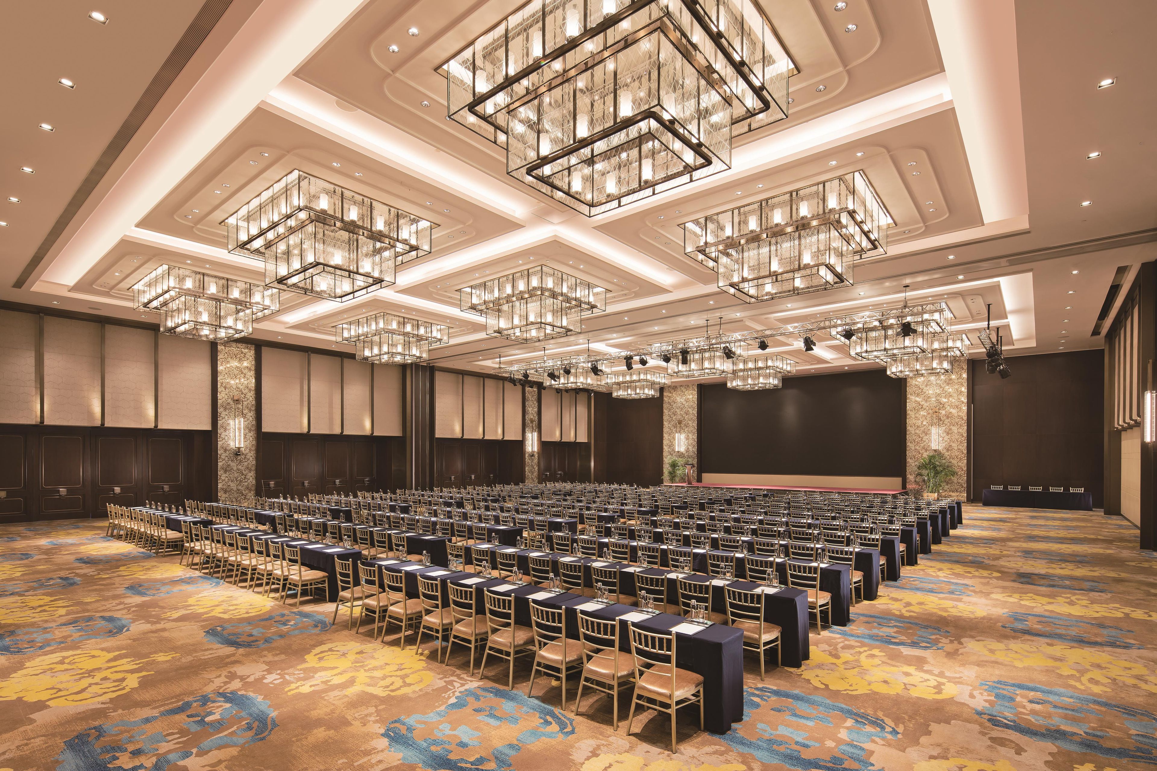 Une grande salle de conférence avec des rangées de tables et de chaises, une scène et un grand écran.