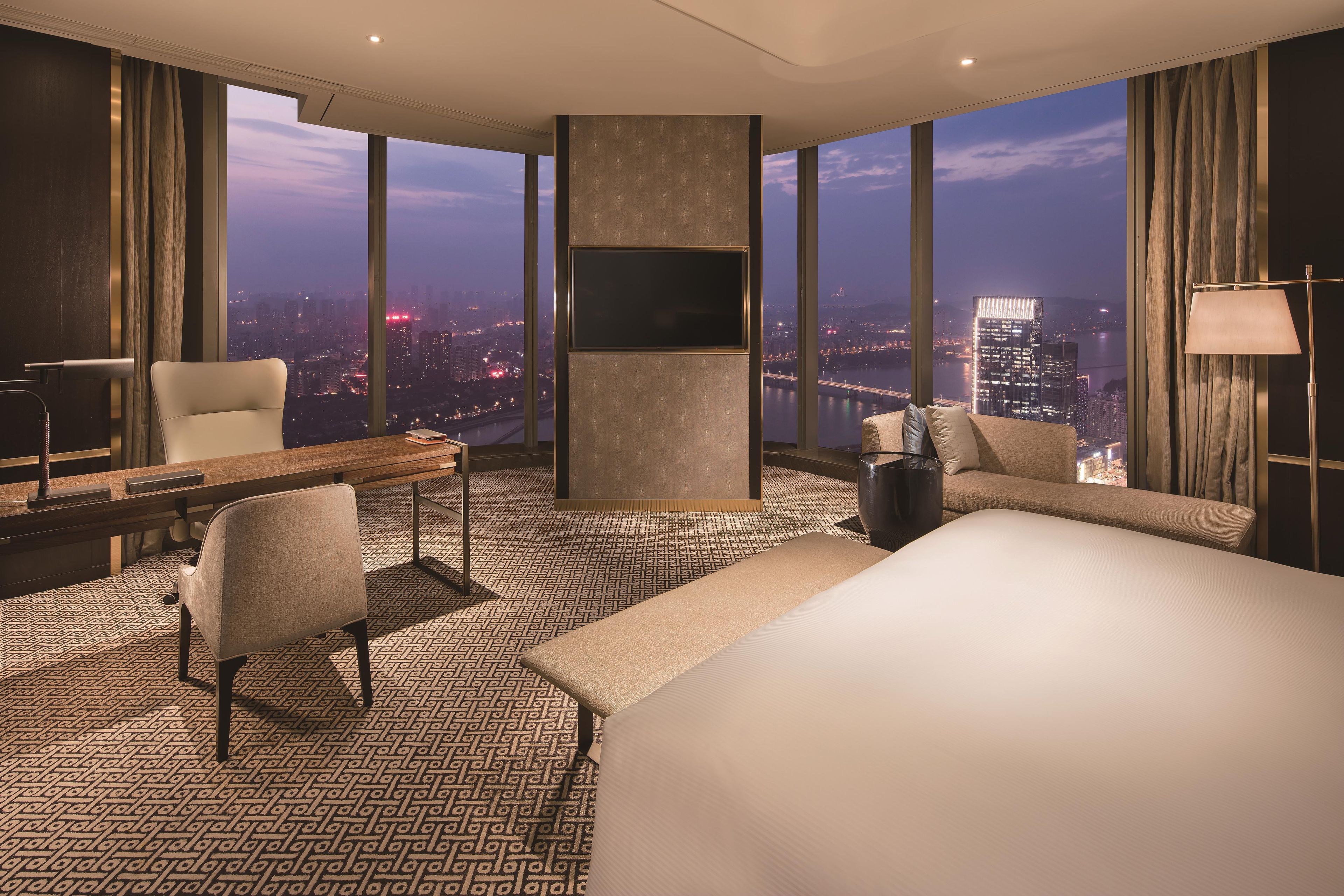 A hotel room with a large bed, a desk, a chair, a TV, and a view of the cityscape.