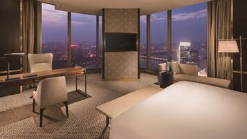 A hotel room with a large bed, a desk, a chair, a TV, and a view of the cityscape.