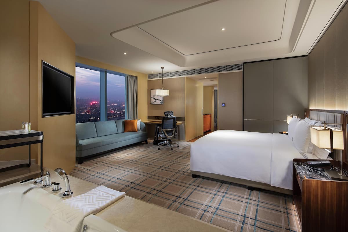 A hotel room with a large bed, a sofa, a desk, a TV, and a view of the city at dusk.