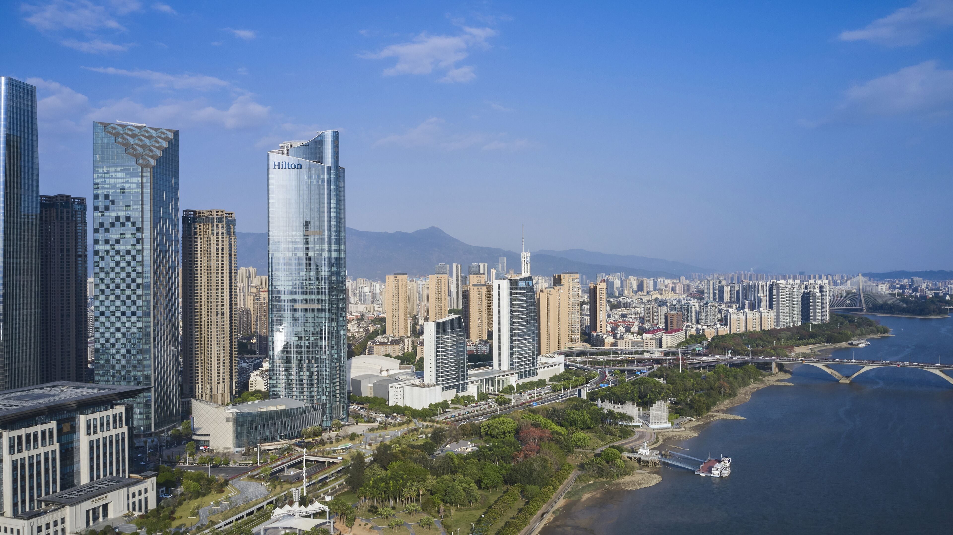 A high-rise hotel with a modern glass facade, located near a river with a bridge in the background.