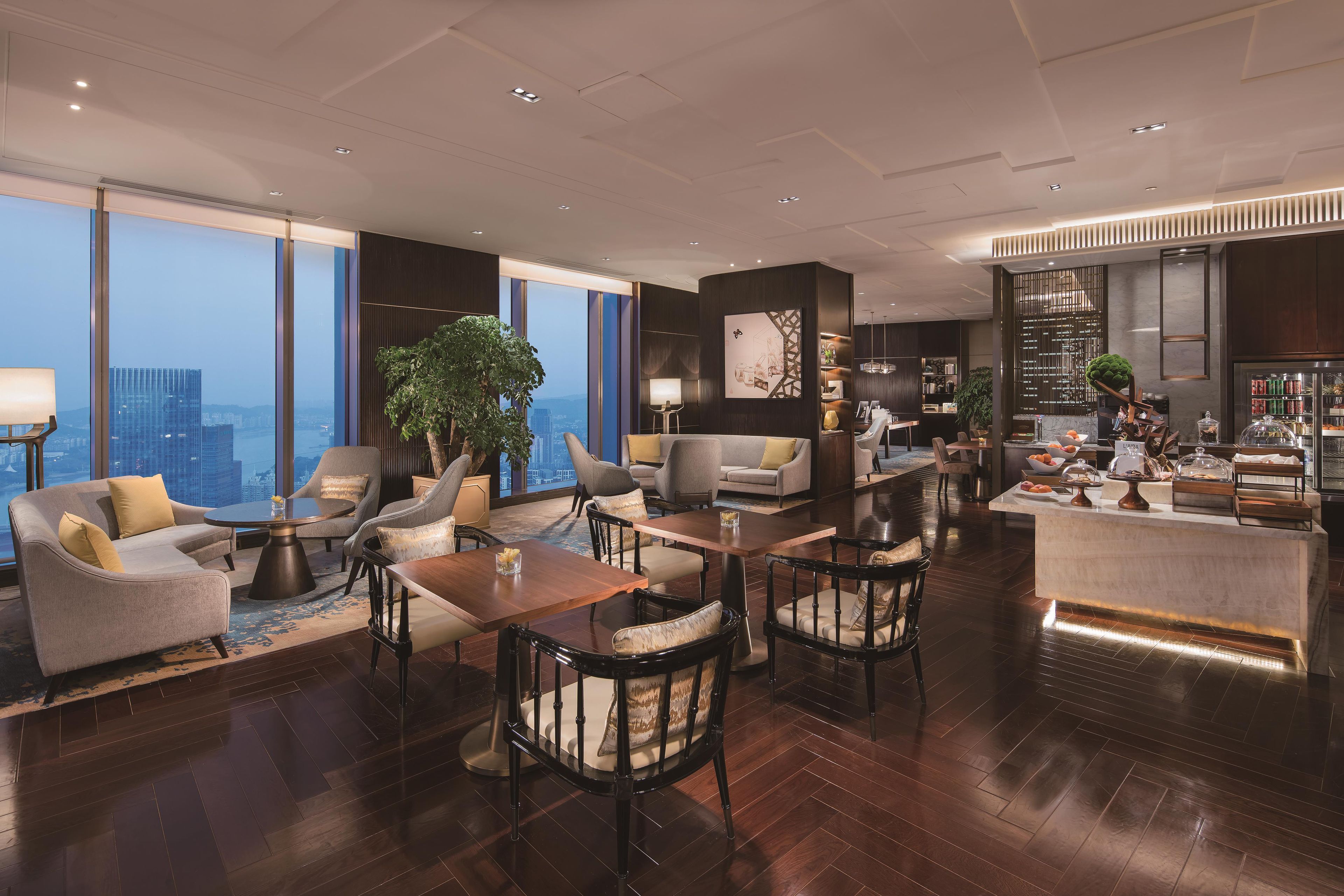 A spacious living area with a high ceiling, large windows offering a city view, and a modern kitchen with a bar area.