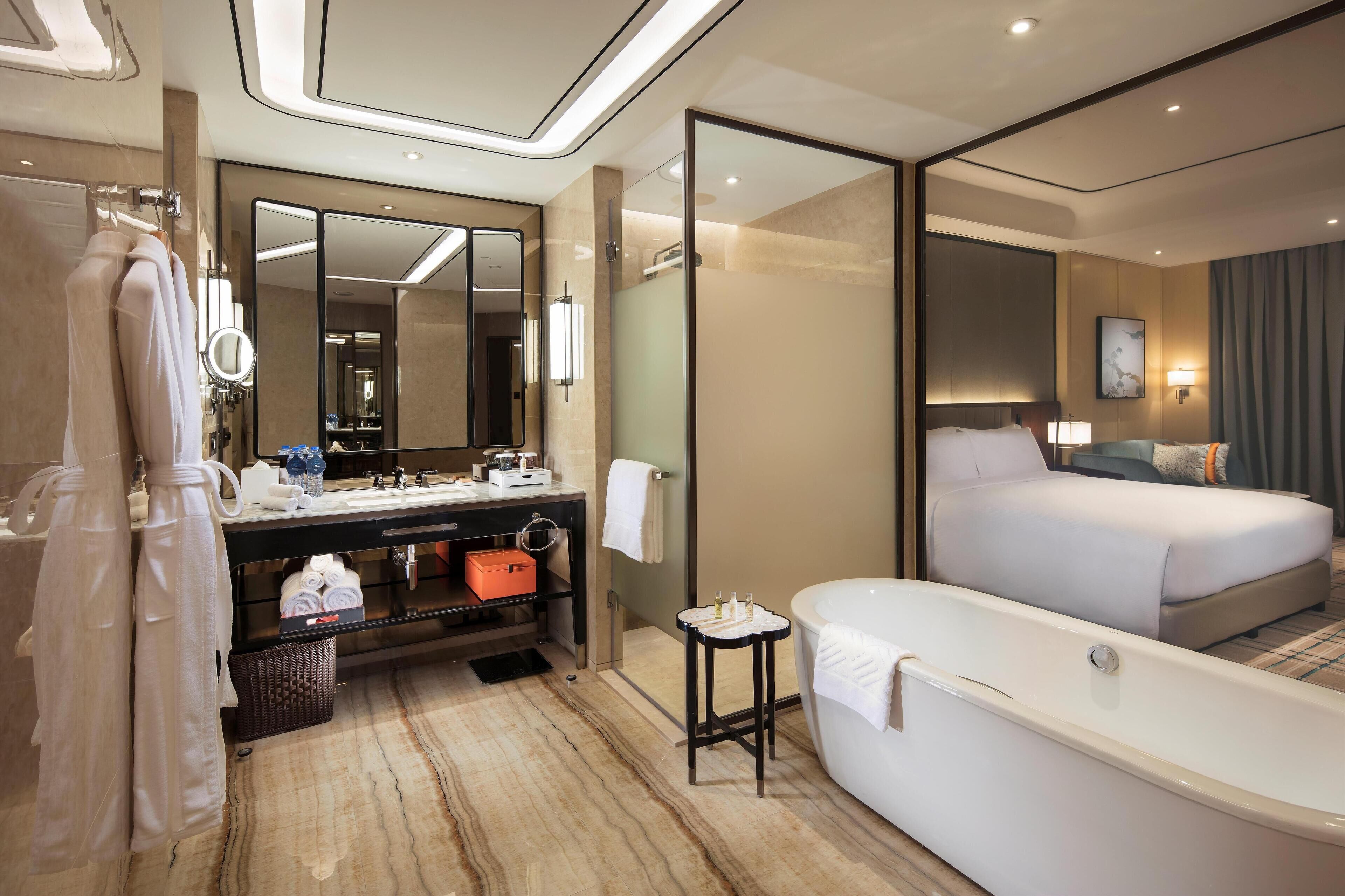 Une chambre d’hôtel moderne dotée d’un grand lit, d’une salle de bain séparée avec une baignoire, d’une coiffeuse avec un miroir et d’un grand miroir qui reflète la pièce.