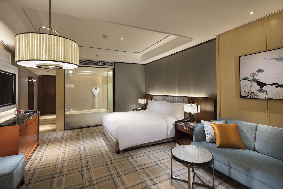 A modern hotel room with a bed, a sofa, a small table, a TV, and a bathroom with a glass partition.
