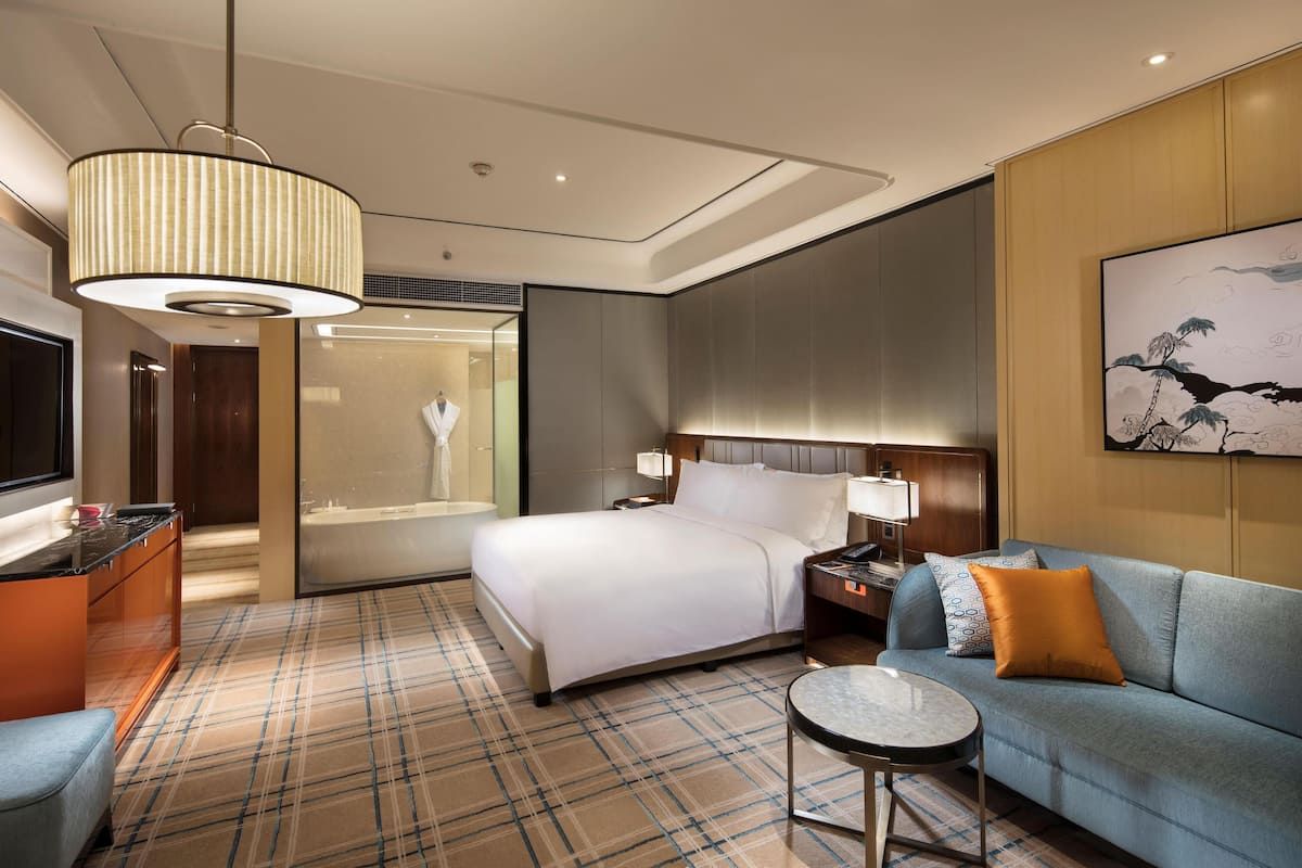 A hotel room with a bed, a sofa, a small table, a TV, and a large painting on the wall.