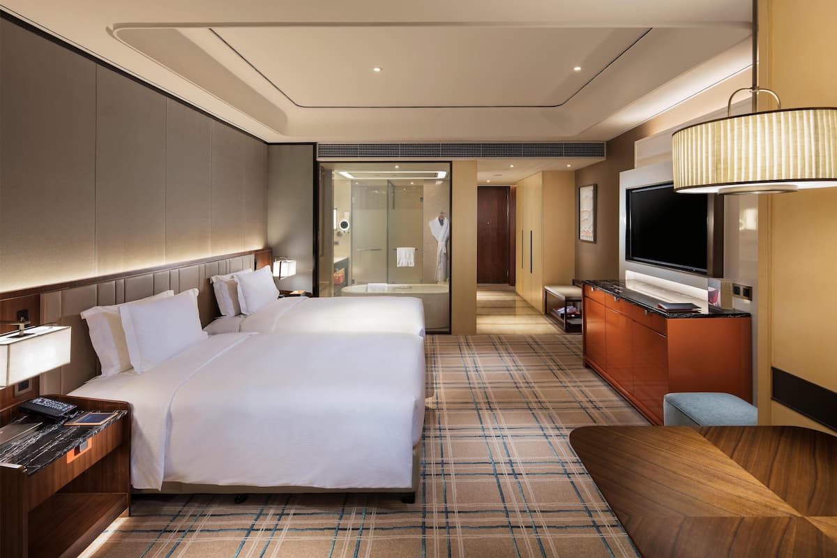 A hotel room with a large bed, a desk, a TV, and a bathroom visible through a glass door.