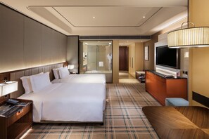 Quarto de hotel com uma cama grande, uma escrivaninha, uma televisão e um banheiro visível através de uma porta de vidro.