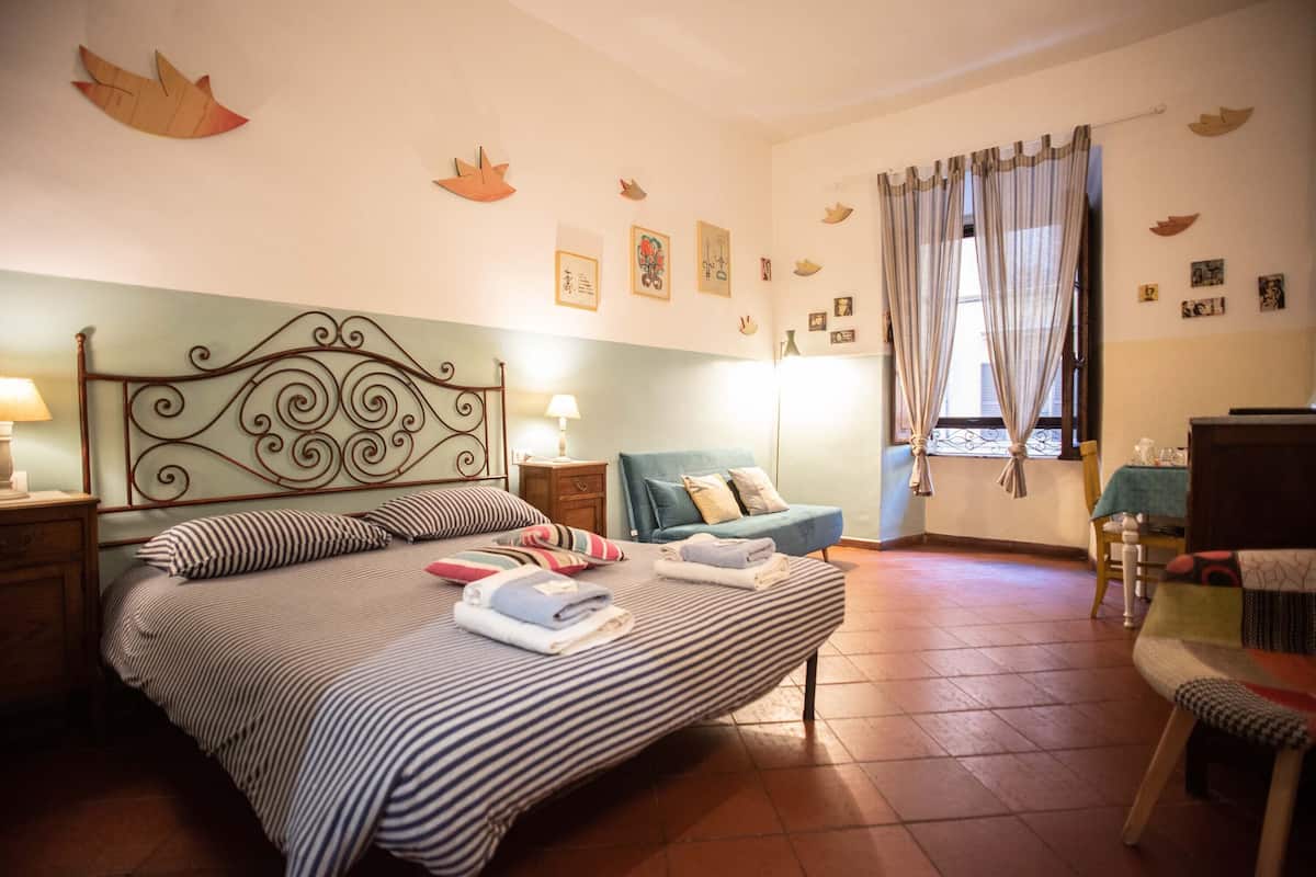 Um quarto com cama, mesas de cabeceira, uma cadeira e uma janela com cortinas.
