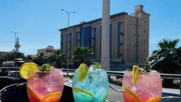 Três cocktails sobre uma mesa, com vista para um edifício alto e céu claro.
