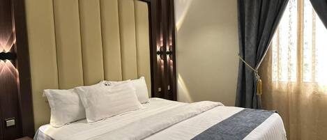 A hotel room with a large bed, a wooden headboard, a bedside table, and a window with curtains.