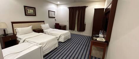 A hotel room with two beds, a bedside table with a lamp, a small table with a phone and water bottles, a mirror, and a window with curtains.