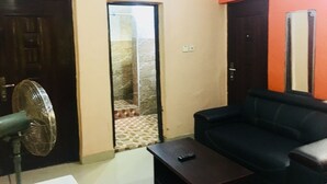 A living room with a black sofa, a small table, and a standing fan. There is a door leading to a hallway with a tiled floor. - HardRock Hotel and suites (Lagos)