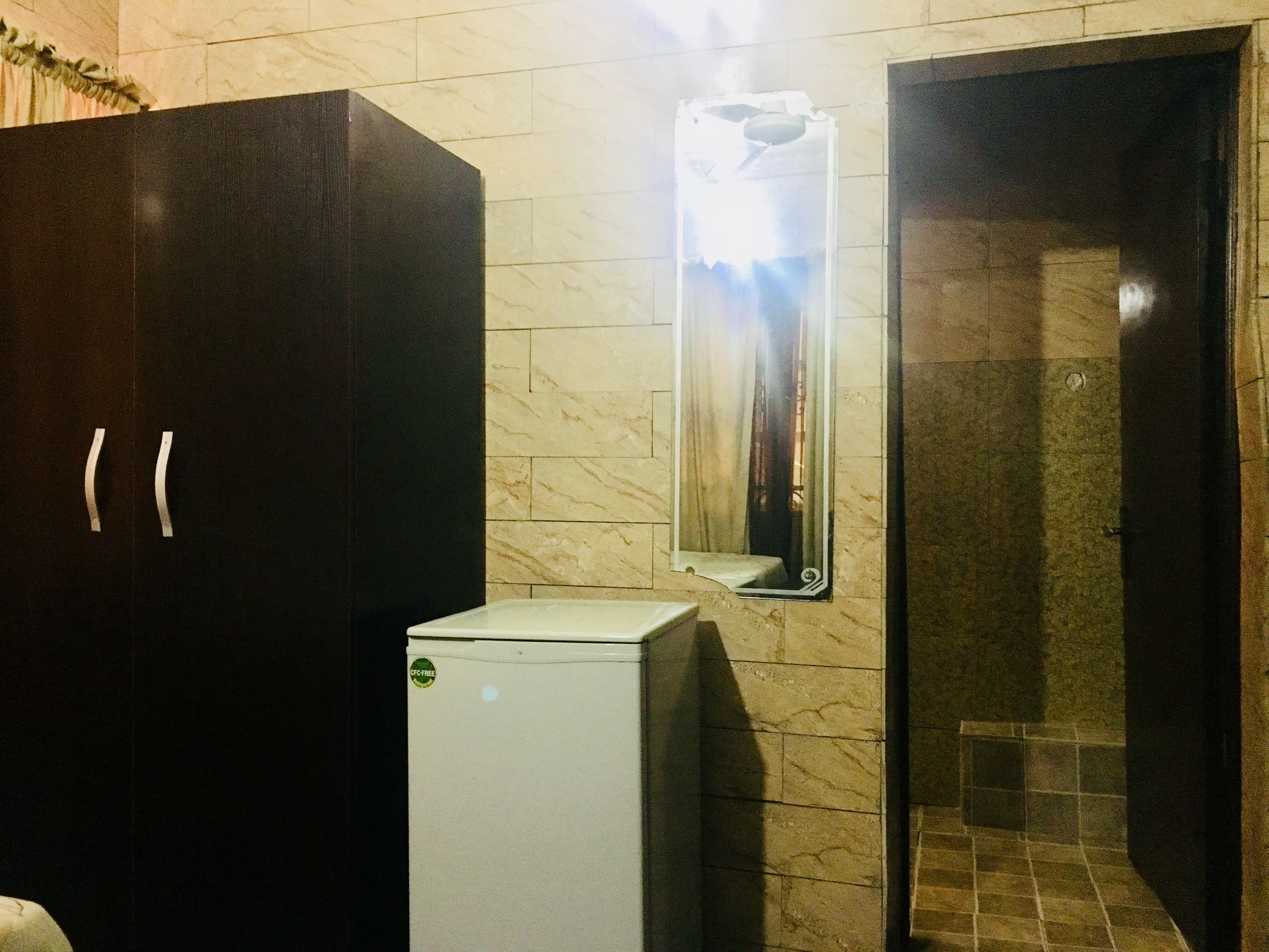 Un baño con un armario de color oscuro, un refrigerador blanco, un espejo con lámpara y una zona de ducha con azulejos.