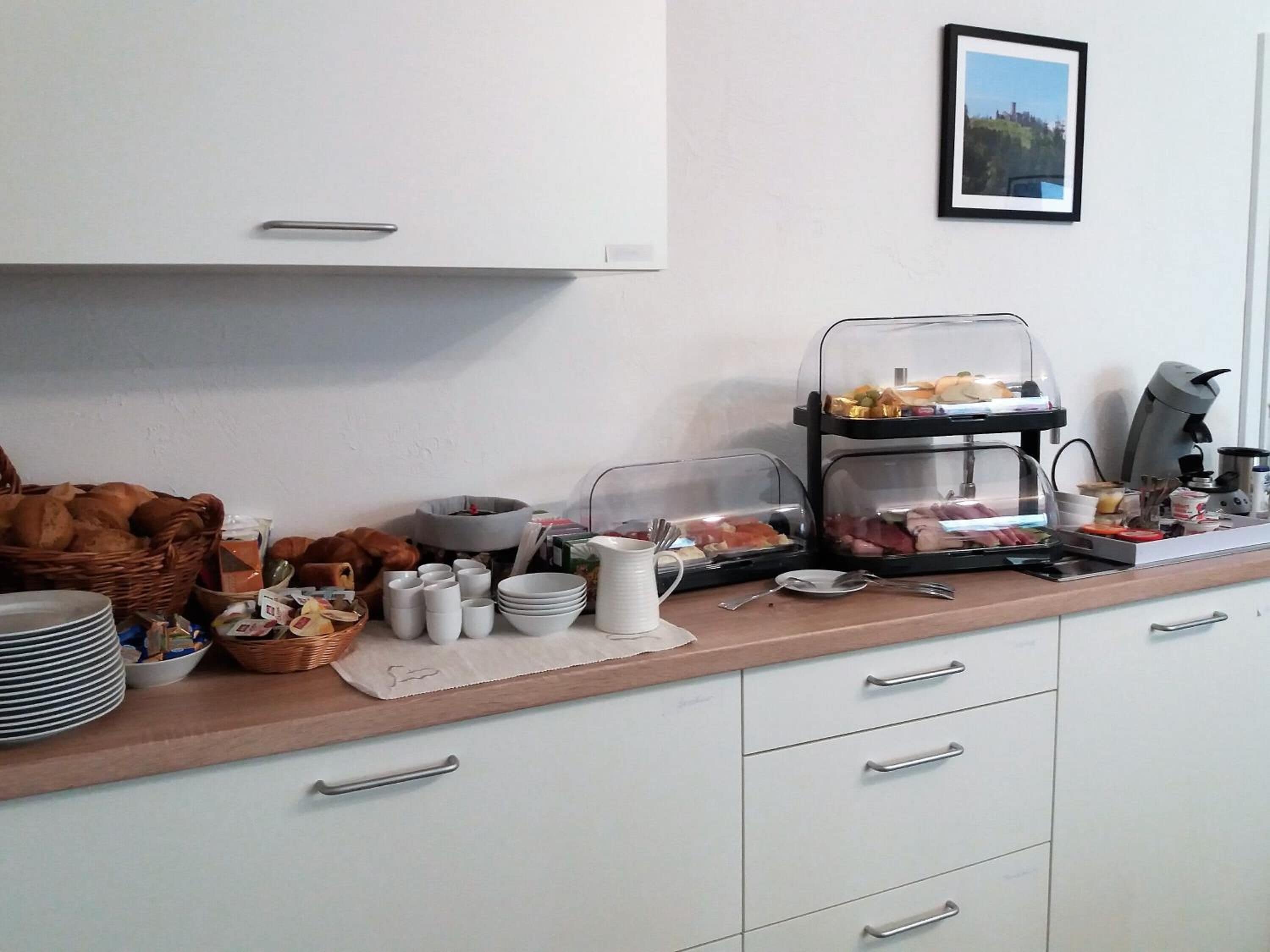 A breakfast buffet setup with various food items and utensils on a counter, accompanied by a coffee machine and a framed picture on the wall.