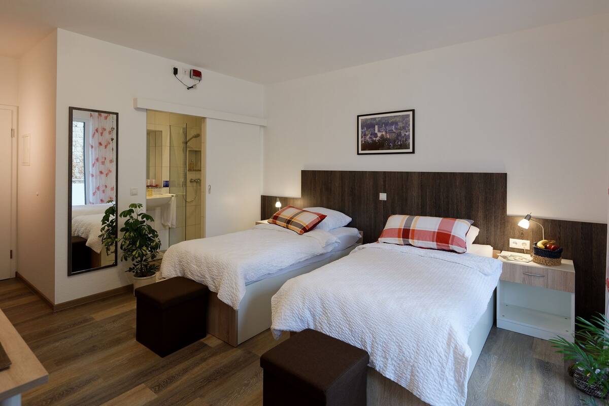 Une chambre d’hôtel avec deux lits, un plancher en bois, une salle de bain avec une douche en verre et une petite table avec une lampe.