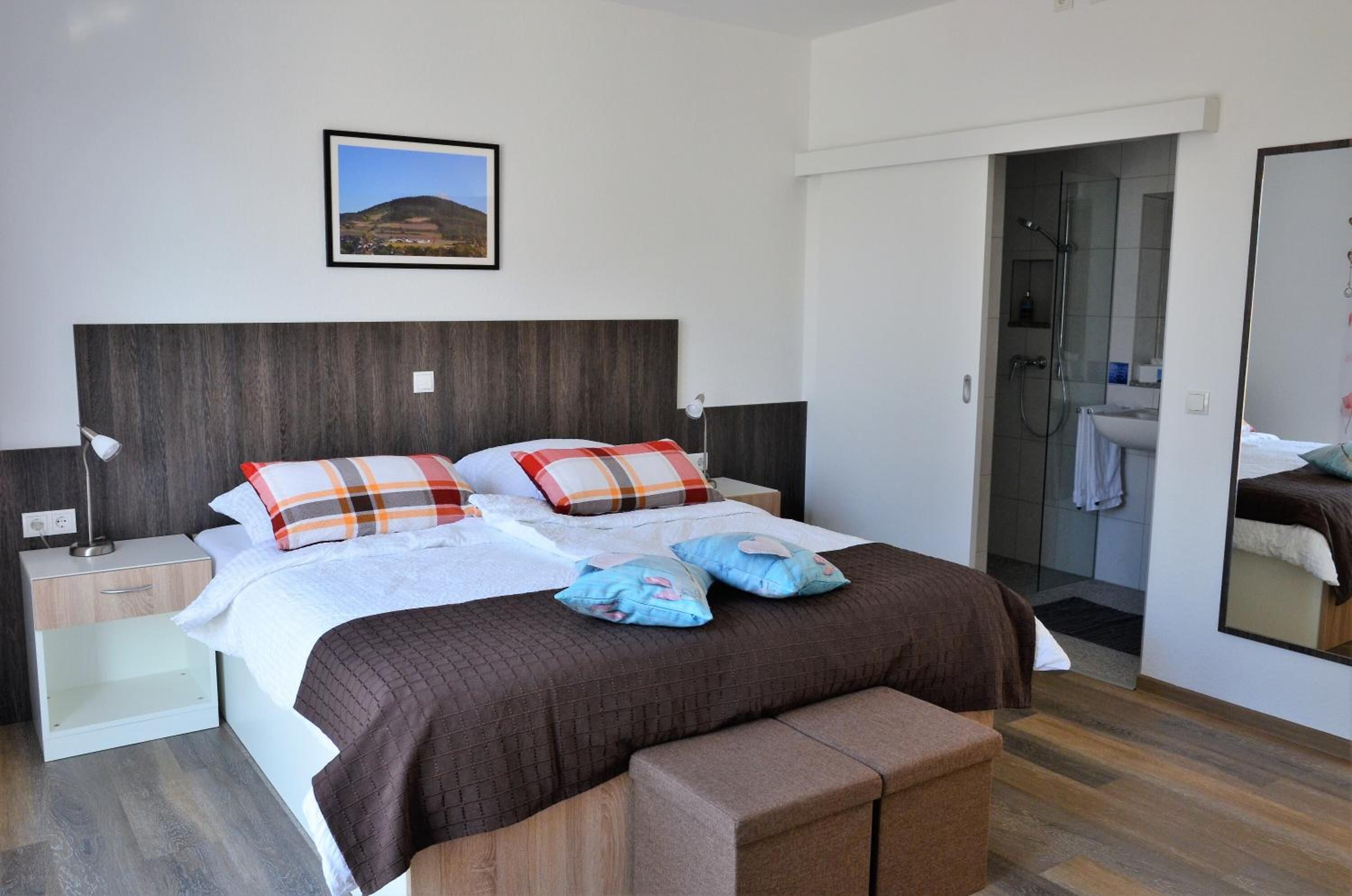 Un dormitorio moderno con una cama grande, una mesita de noche, un tocador, un espejo y un baño visible a través de una puerta abierta.
