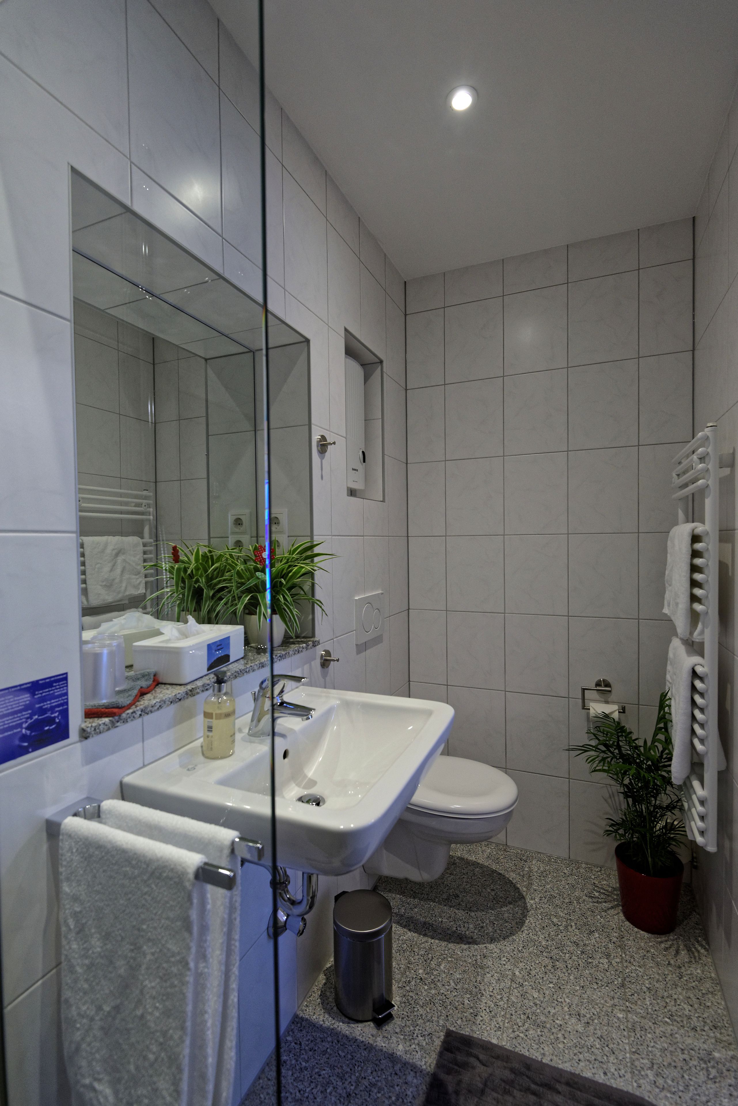 A modern bathroom with a large mirror, white tiles, a sink, a toilet, and a potted plant.