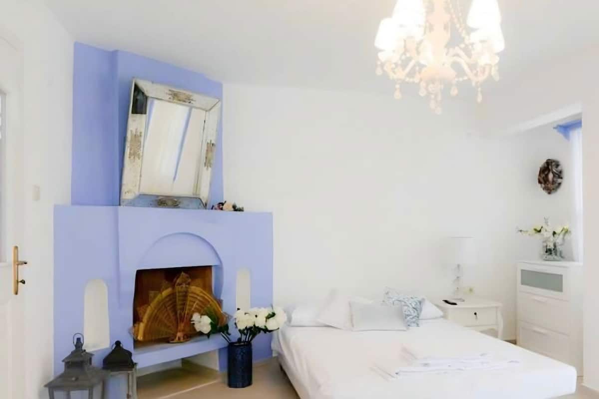 A bedroom with a white bed, a blue accent wall, a fireplace, and a chandelier.