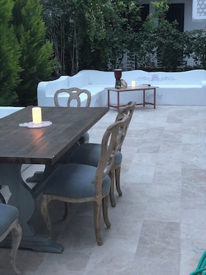 An outdoor patio with a wooden table and chairs, a white cushioned bench, and a small table with a candle. - Kiraz Cicegi Butik Akyaka (Ula)