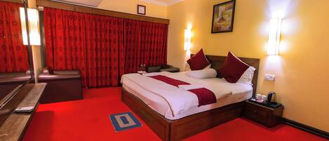 A hotel room with a large bed, red curtains, a bedside table with a lamp, and a nightstand with a lamp.