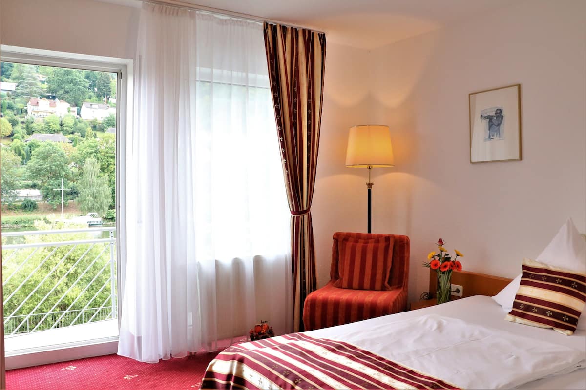 A hotel room with a bed, a chair, a lamp, a painting, and a window with curtains.