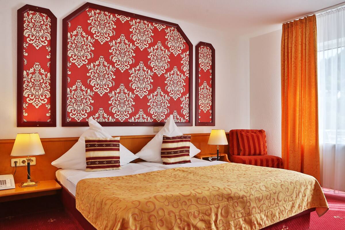 A hotel room with a large bed, two bedside tables, a nightstand, a red chair, and a window with curtains.
