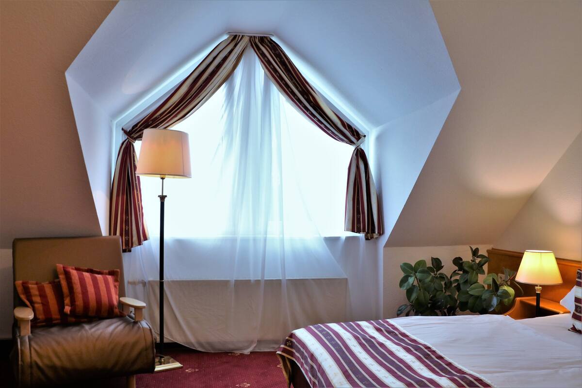 A hotel room with a large window, a bed, a chair, a floor lamp, and a potted plant.