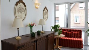 A room with a wooden desk, two lamps, a red sofa, and a large window with a view of a brick building. - Hotel Neckarlux (Heidelberg)