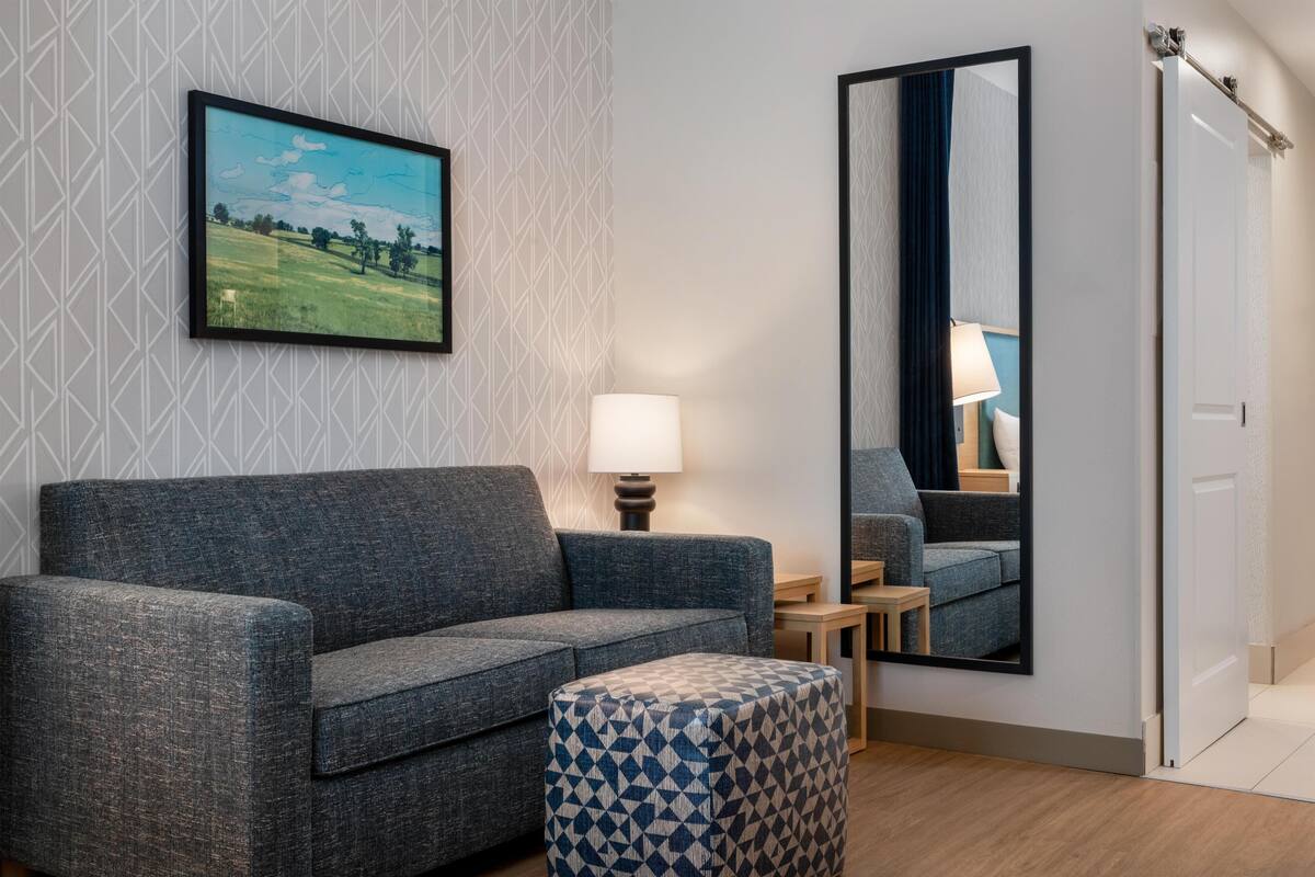 Une pièce avec un canapé gris, un pouf à motifs, une lampe, un miroir et un tableau encadré accroché au mur.