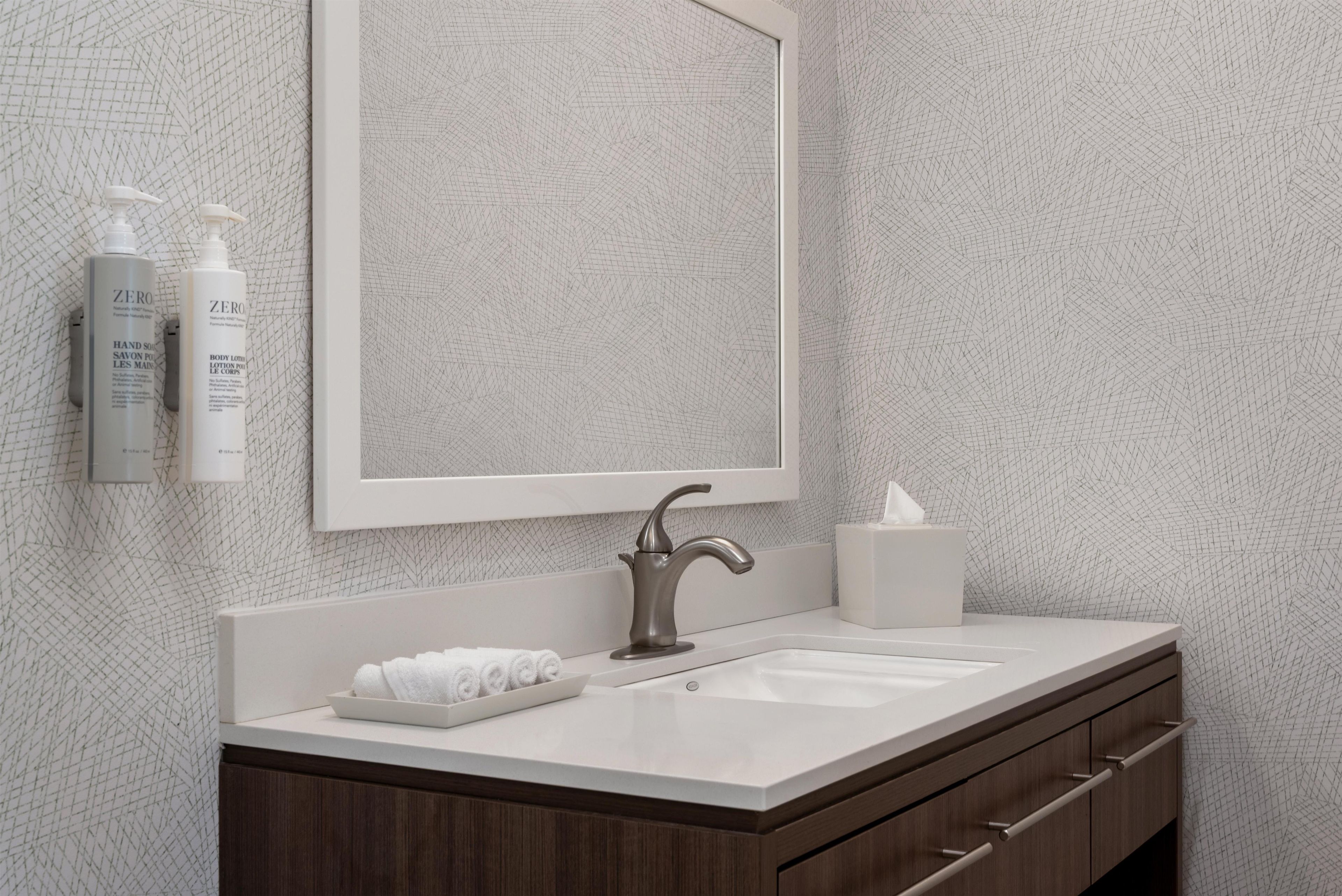 Un baño con lavamanos blanco, tocador de madera oscura, un espejo grande y dos botellas de jabón para manos.