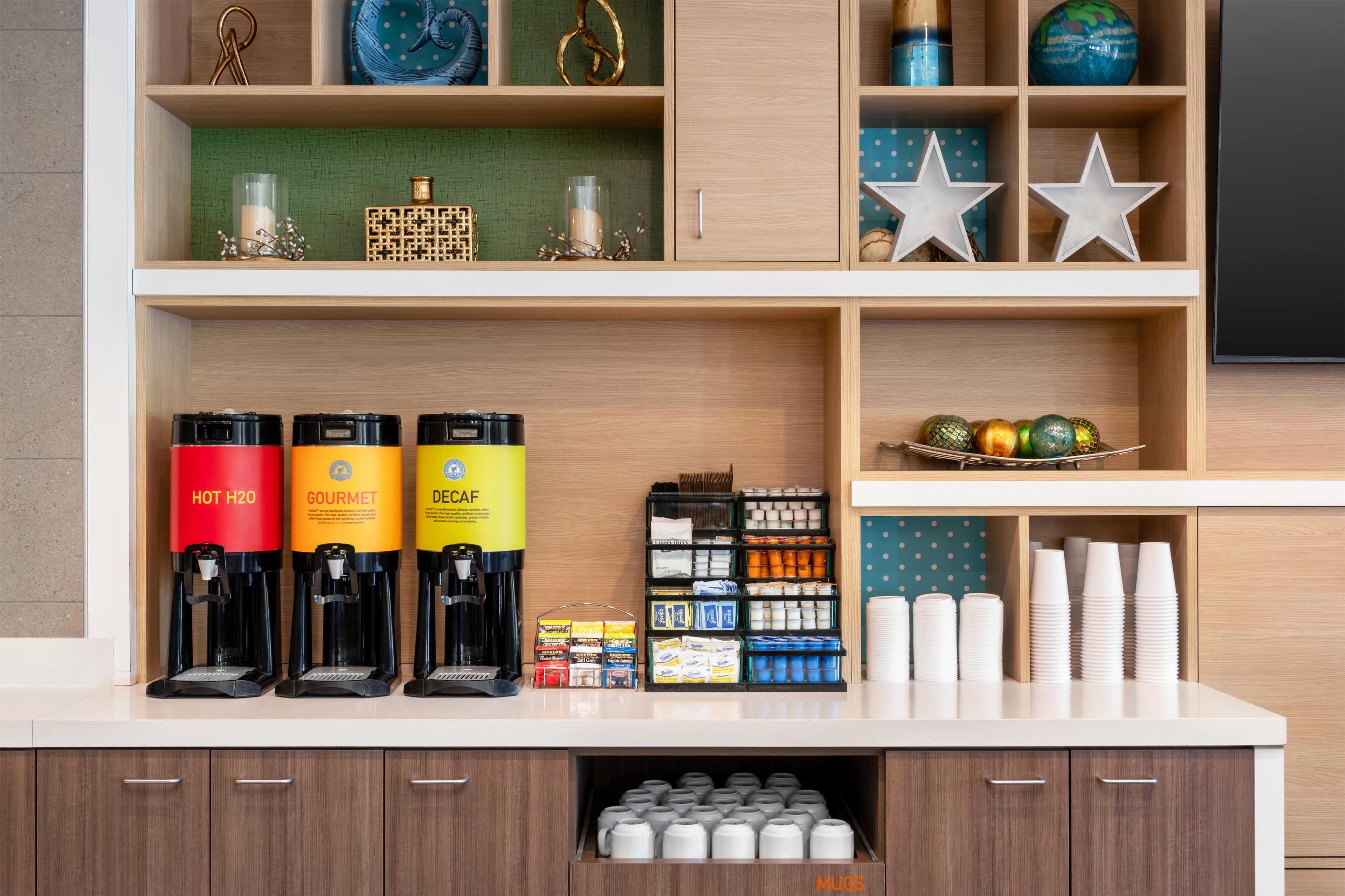 Eine Kaffeestation mit drei Kaffeeautomaten, beschriftet mit 'HOT H2O', 'GOURMET' und 'DECAF'. Außerdem sind Regale mit verschiedenen Gegenständen zu sehen, darunter ein Globus, dekorative Sterne und ein Korb voller farbenfroher Ornamente.