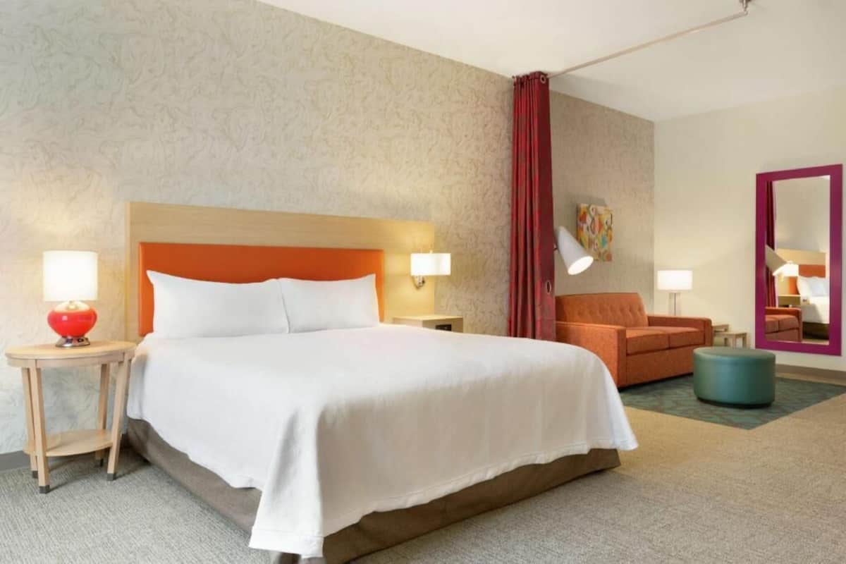 Une chambre d’hôtel avec un grand lit, deux tables de chevet avec des lampes, un canapé, un miroir et un sol recouvert de moquette.