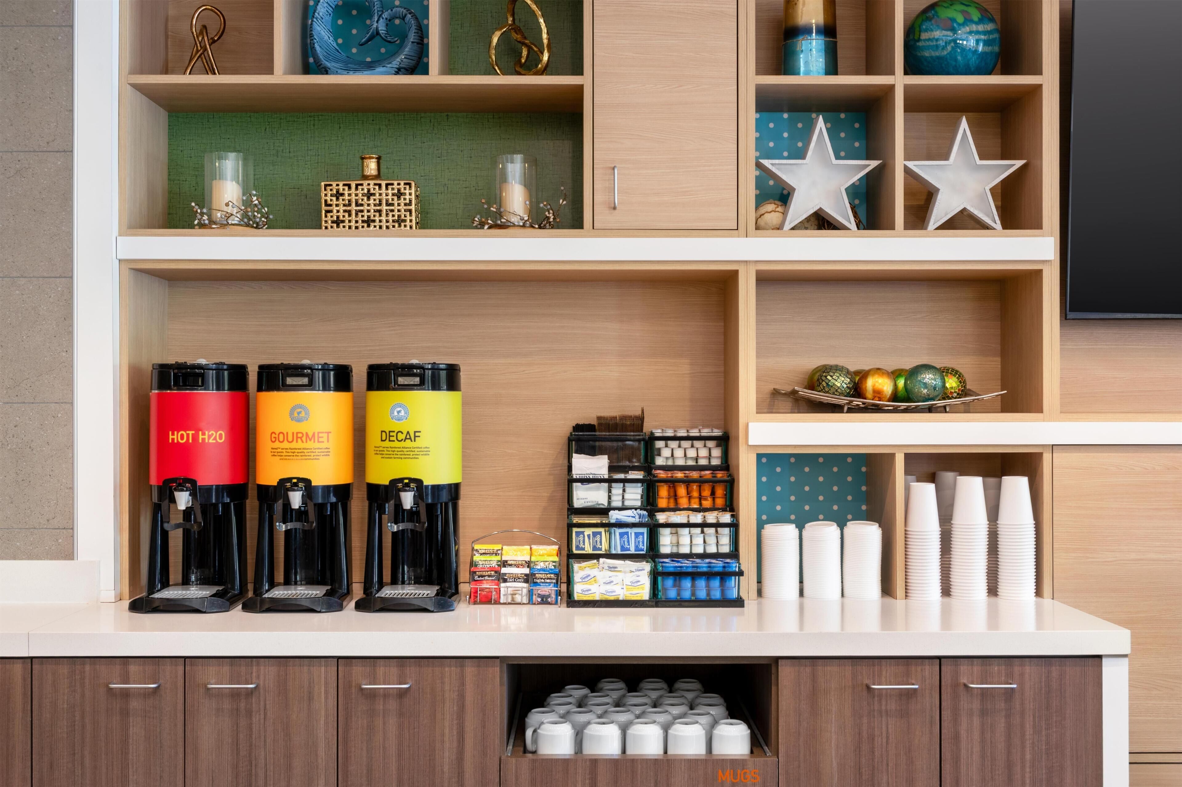 Een koffiehoek met drie koffiemachines, genaamd 'HOT H2O', 'GOURMET' en 'DECAF'. Er zijn ook planken met diverse artikelen, waaronder een wereldbol, decoratieve sterren en een mand met kleurrijke versieringen.