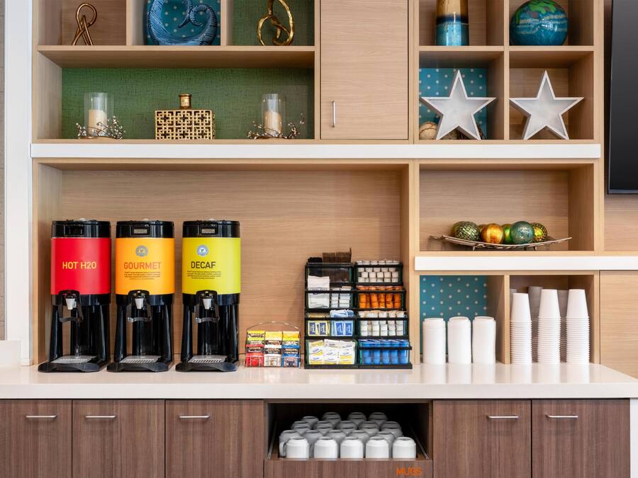 A coffee station with three coffee machines labeled 'HOT H2O,' 'GOURMET,' and 'DECAF.' There are also shelves with various items, including a globe, decorative stars, and a basket of colorful ornaments.