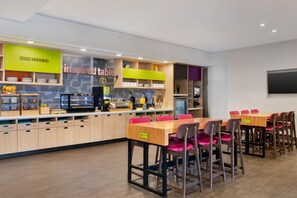 A modern cafe with a counter displaying breakfast items, a 'Good Morning' sign, and a 'Inspired Table' sign. There are wooden tables and chairs with a clean, minimalist design.