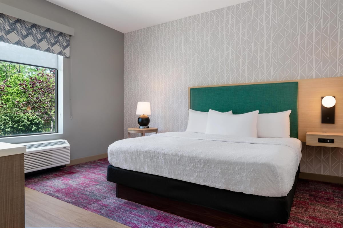 Une chambre d’hôtel avec un lit, une table de chevet avec une lampe, un luminaire fixé au mur et une fenêtre avec un climatiseur.