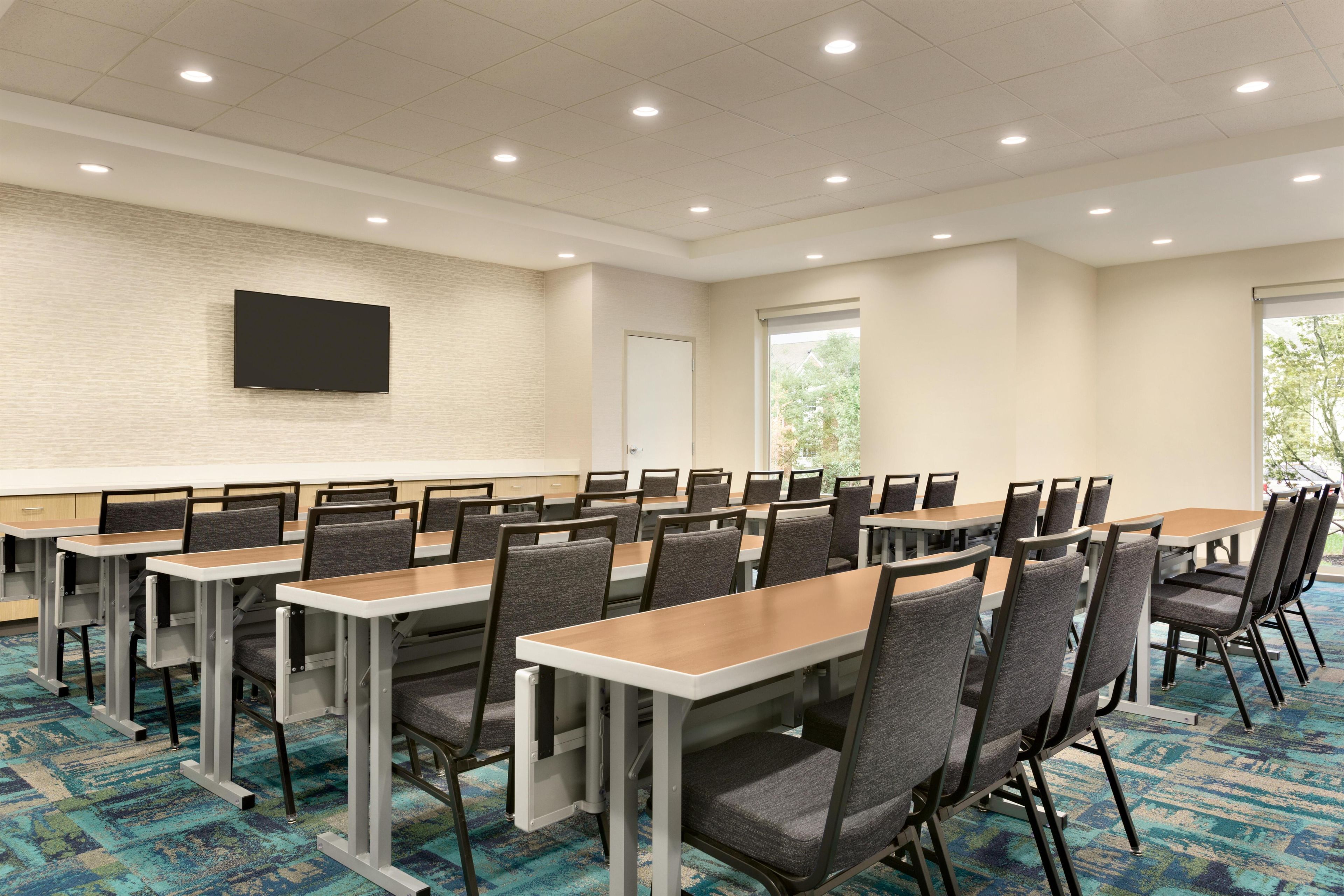 A conference room with tables and chairs, a flat-screen TV on the wall, and windows with a view of trees.