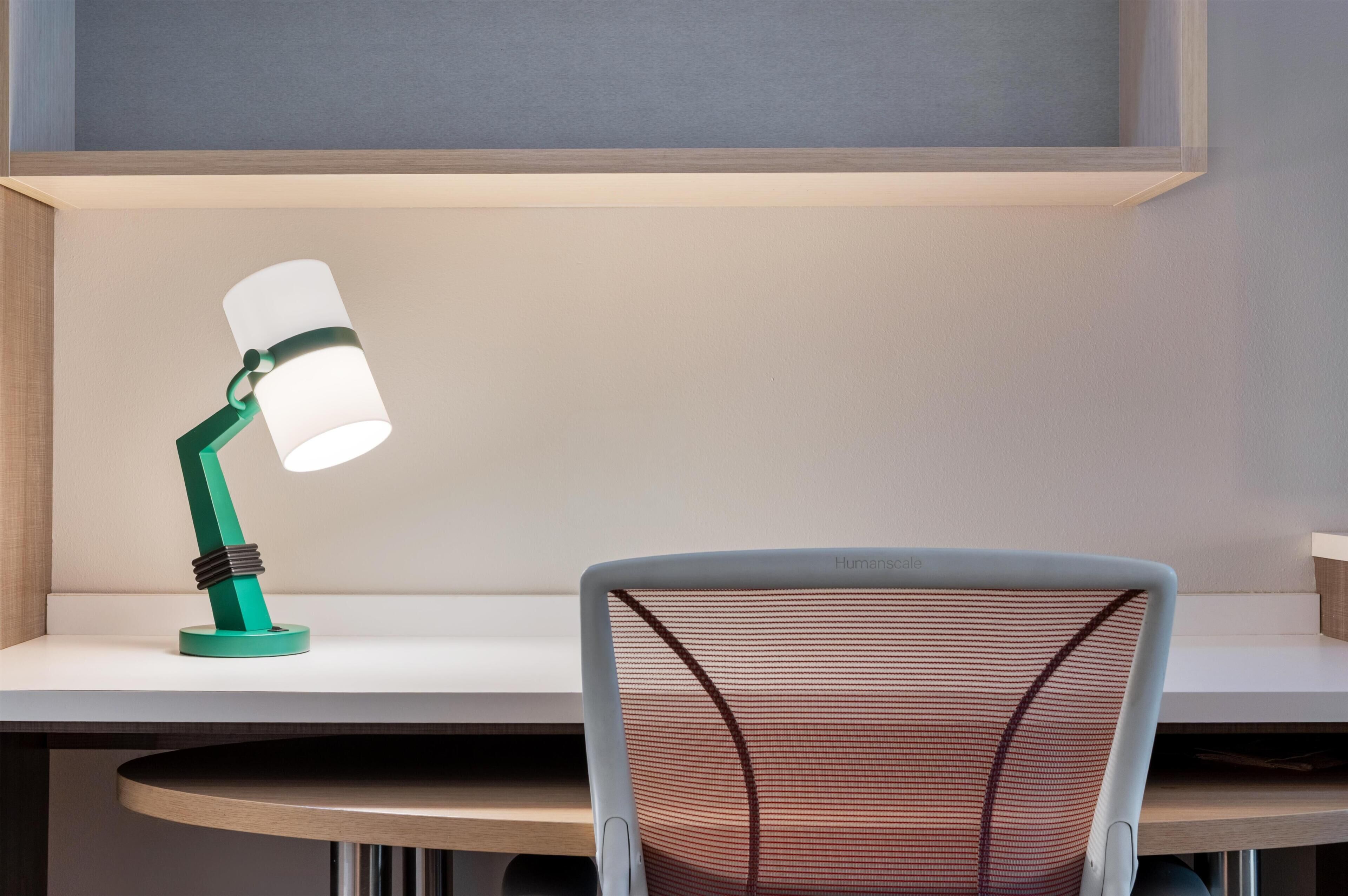 A desk with a green desk lamp, a white chair, and a shelf above.
