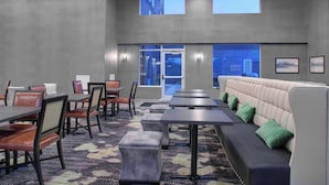 Un moderno hall de hotel con una larga mesa de comedor, asientos acolchados y grandes ventanales con vistas a la ciudad.