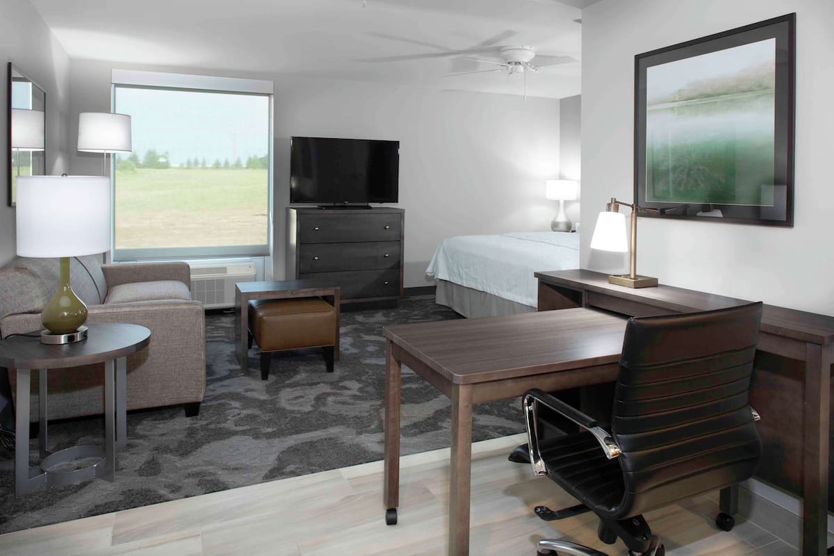 Ein Hotelzimmer mit Bett, Schreibtisch, Stuhl, Fernseher, Lampe und einem großen Fenster mit Blick auf ein Feld.
