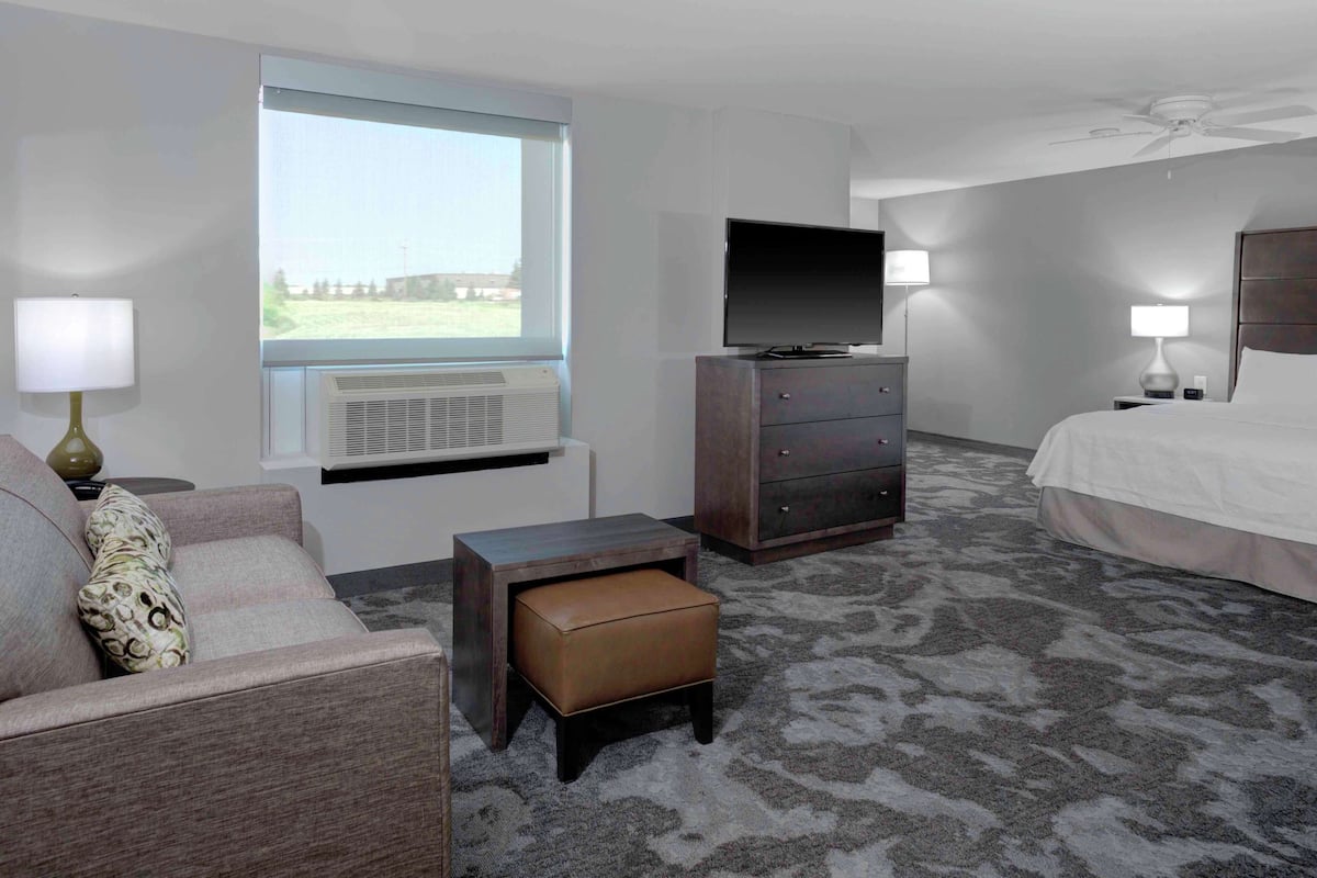 Ein Hotelzimmer mit Bett, Fernseher, Kommode, einem kleinen Tisch, einer Couch und einem Fenster mit Klimaanlage.