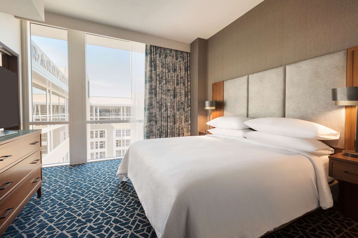 A hotel room with a large bed, a dresser, a nightstand with a lamp, and a view of the city through large windows.