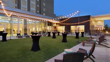 An outdoor patio area with lounge chairs and tables, a pool, and a building with large windows and string lights.