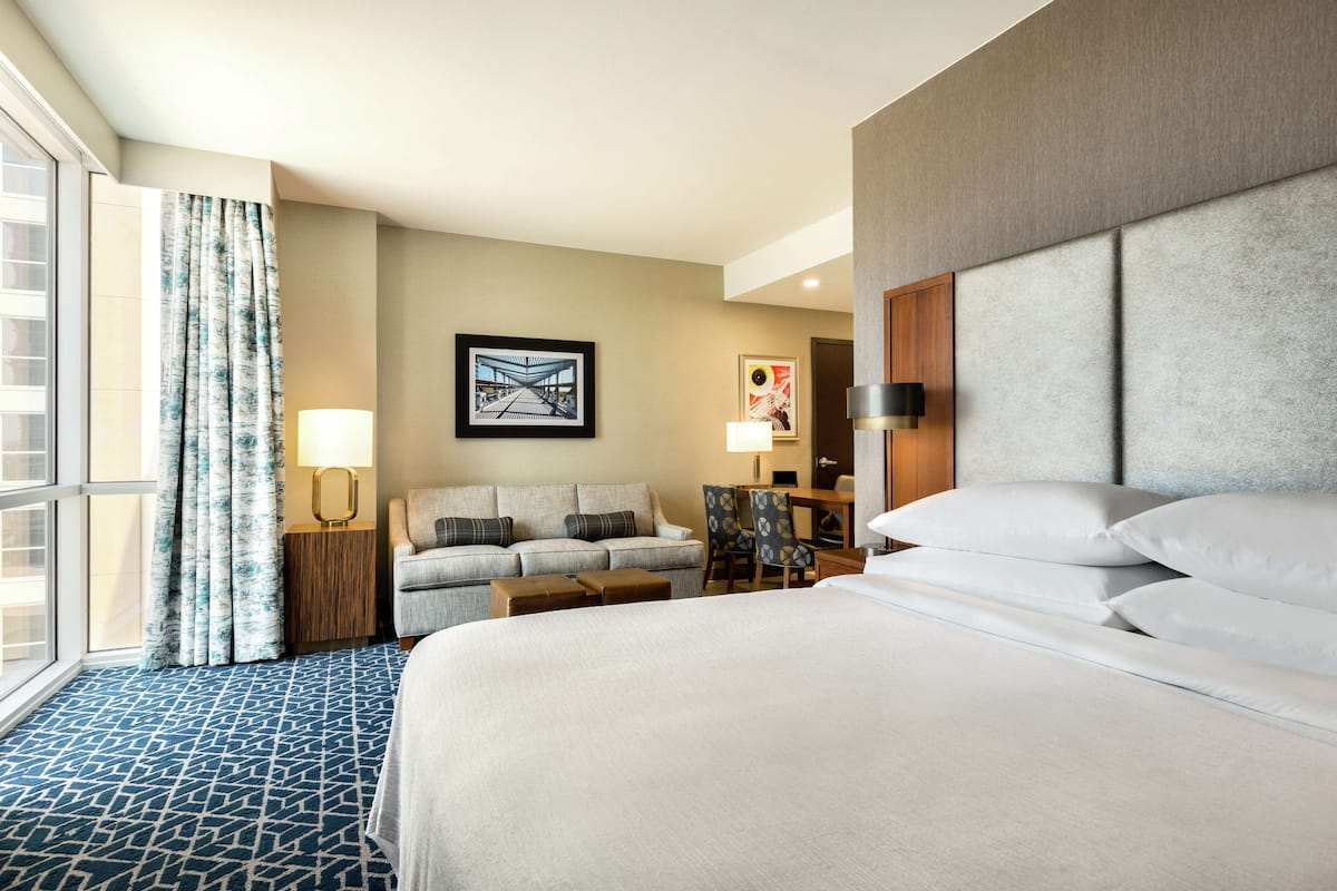 A hotel room with a large bed, a sofa, a desk, and a chair. There are two lamps, a window with curtains, and artwork on the wall.