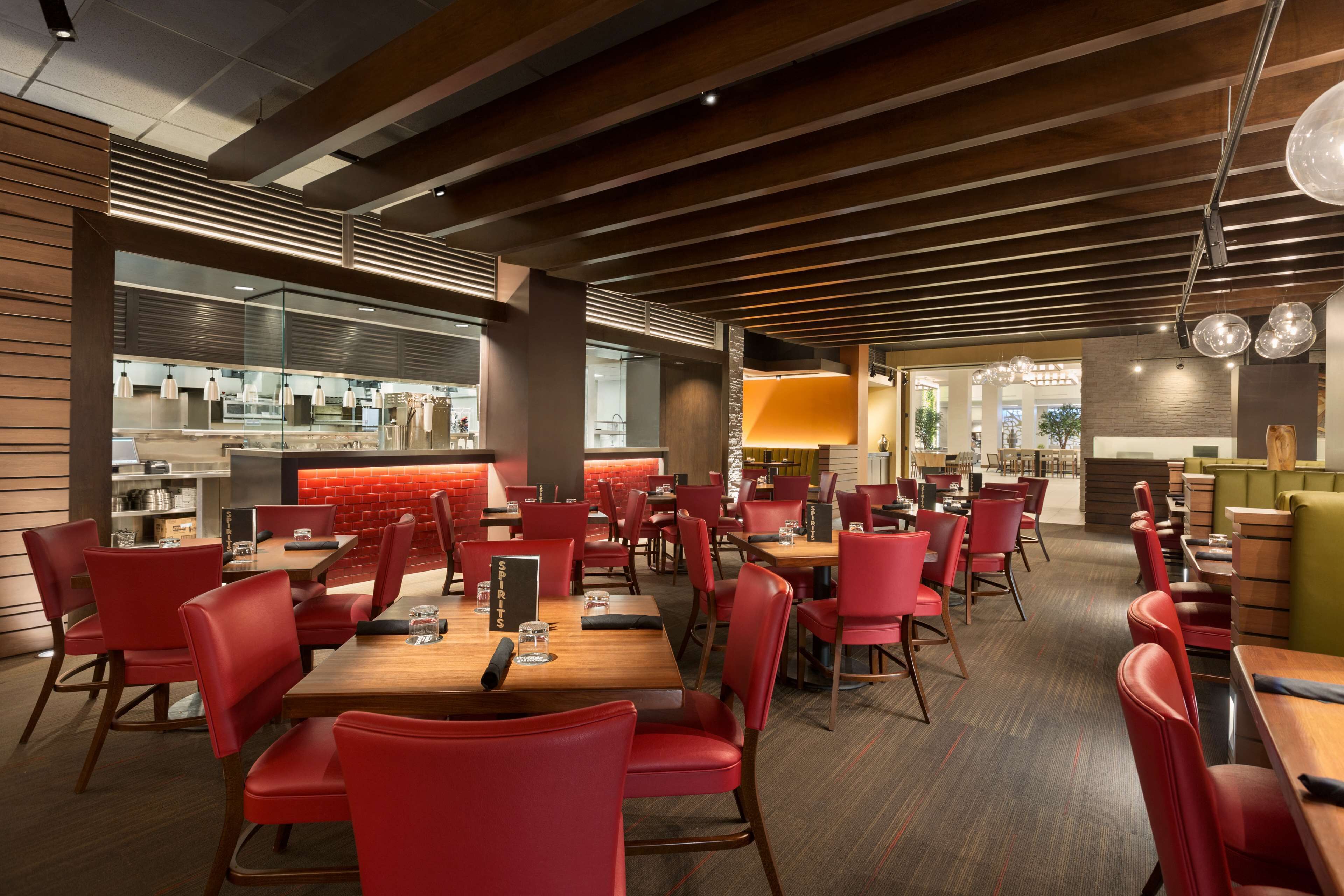 A modern restaurant with red chairs and wooden tables, featuring a kitchen area in the background.