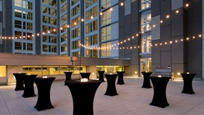 A courtyard with multiple tables and chairs, string lights, and a building with large windows in the background. - Embassy Suites by Hilton Denton Convention Center (Denton)